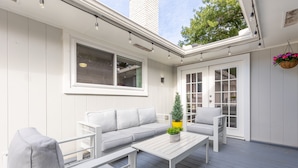 A covered patio with a white sofa, a white table, and a hanging planter with flowers. - Private Family Heated Pool and Spa, 2 King Beds, Cornhole, Grill, Foosball, Game (Richardson)