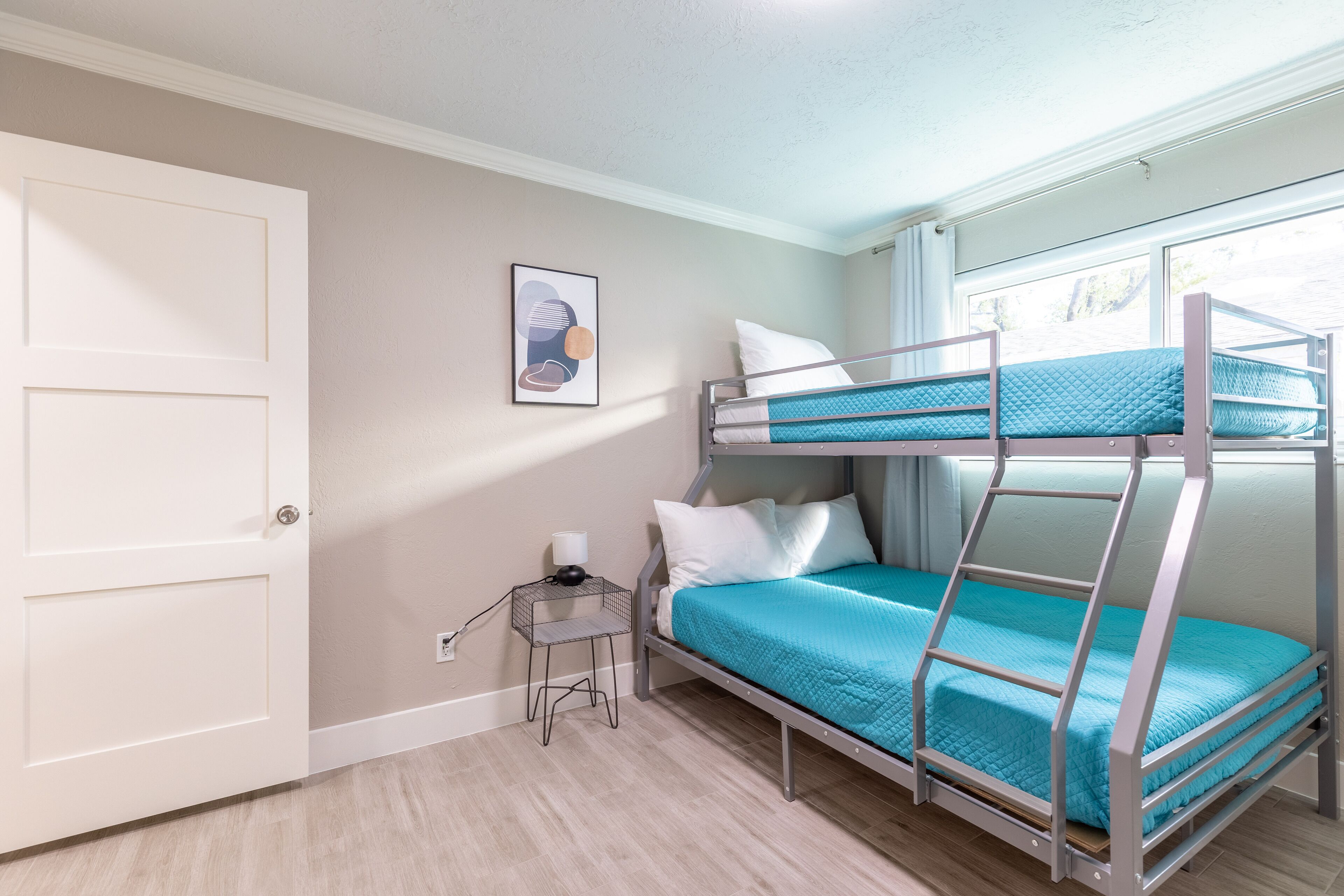 A bunk bed room with a white door, a small bedside table with a lamp, and a framed picture on the wall.