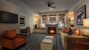 A cozy living room with a fireplace, a ceiling fan, and a large window with a view of trees.