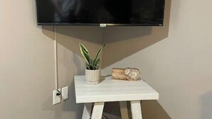 Seinälle asennettu litteänäyttöinen televisio, sen alla pieni valkoinen pöytä, jonka päällä on kasvi ja koriste-esine.