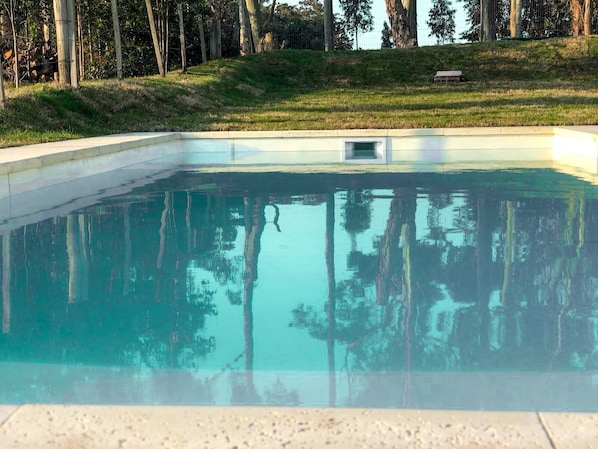 Piscina retangular com água cristalina refletindo árvores e uma área gramada.