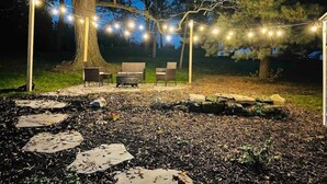 Un espace extérieur aménagé avec des guirlandes lumineuses, un grand arbre et un foyer en pierre.