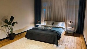 Ein Schlafzimmer mit Bett, Nachttischen, einer Topfpflanze und einem Fenster mit Vorhängen.