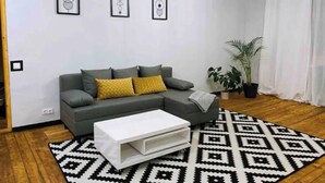 Ein modernes Wohnzimmer mit grauem Sofa, gelben Kissen, einem weißen Couchtisch und einem schwarz-weiß gemusterten Teppich. An der Wand hängen drei gerahmte Kunstwerke und ein Blumentopf steht dort.