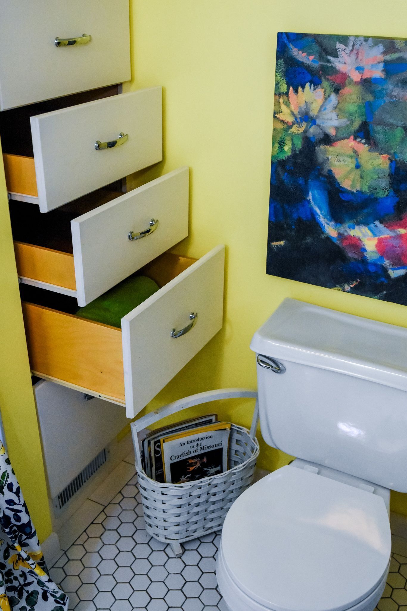 A bathroom with a white toilet, a white basket with books, and a colorful abstract painting on the wall.