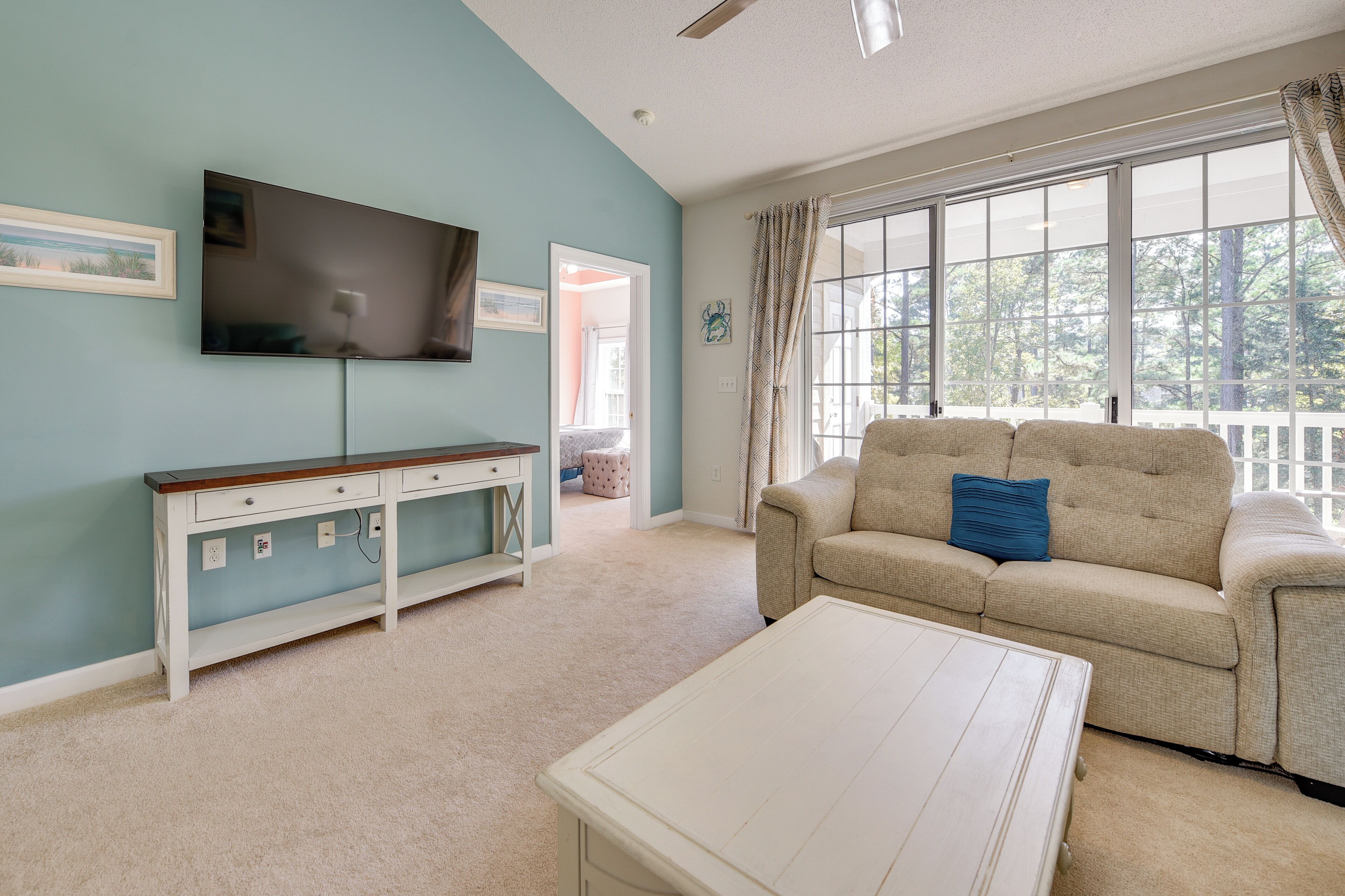 Un salon avec un canapé beige, une table basse blanche, un téléviseur fixé au mur et une grande fenêtre avec des rideaux.