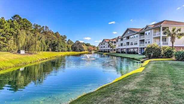 A hotel with a swimming pool, surrounded by greenery and a clear sky. - Pool Access + Balcony: Barefoot Resort Retreat! (North Myrtle Beach)