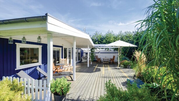 Eine überdachte Terrasse im Freien mit Holzboden, weißem Zaun und Pflanzen in Töpfen.