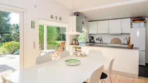 Eine moderne Küche mit weißen Schränken, ein Essbereich mit weißem Tisch und Stühlen sowie ein Blick auf einen Garten durch große Fenster.