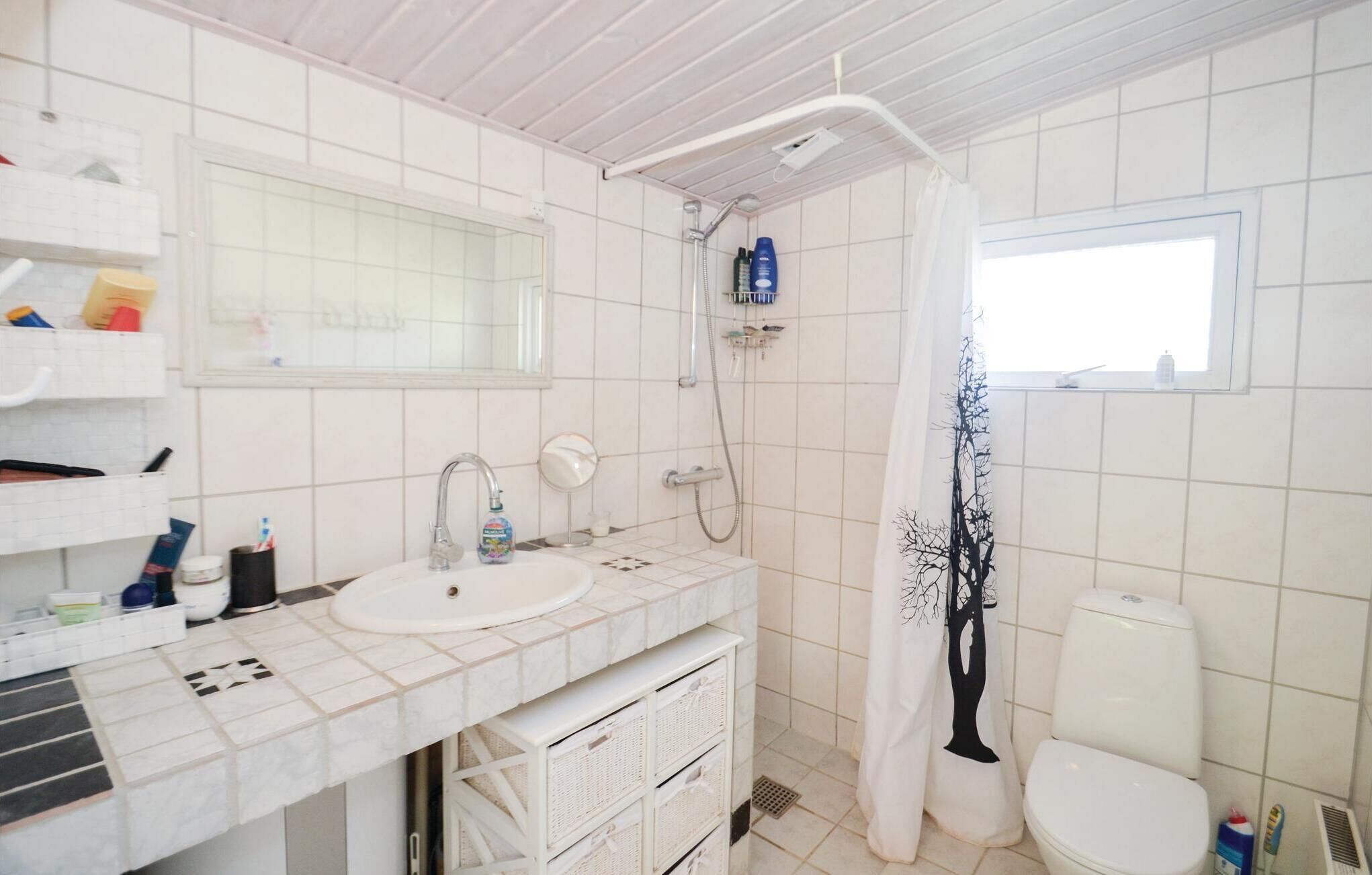 Ein Badezimmer mit weißem Waschbecken, Spiegel und Toilette. Eine Dusche mit Duschvorhang ist vorhanden, auf dem Waschtisch liegen Toilettenartikel.