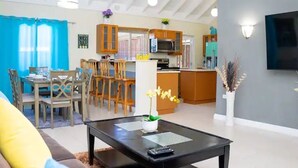 Una sala de estar moderna con un sofá, una mesa de centro y un comedor con sillas y una mesa. Se observa una zona de cocina con alacenas y un microondas.