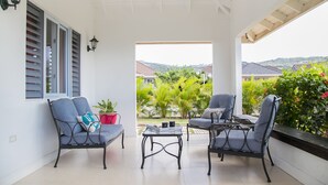 Un patio techado con dos sillas y una mesa, vistas a zonas verdes y casas, y un ventilador de techo.