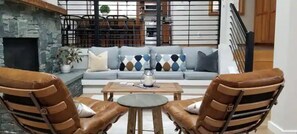 Un salon doté d’une cheminée en pierre, d’un canapé gris agrémenté de coussins à motifs et de deux fauteuils en cuir, avec une table basse ronde placée au centre.
