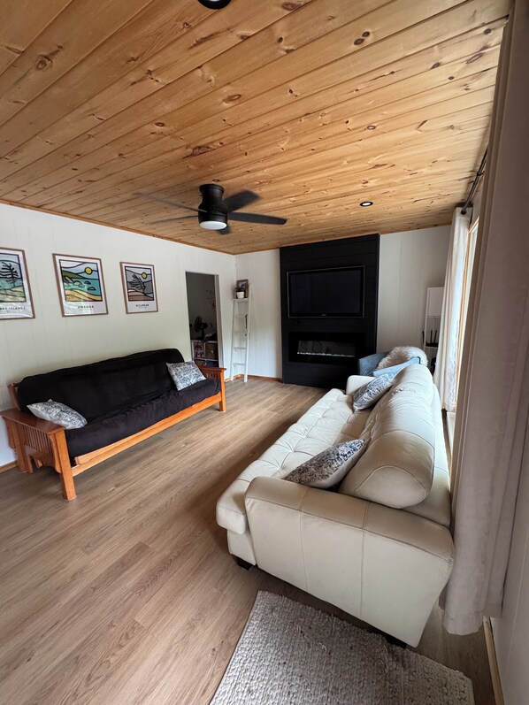 A living room with a wooden ceiling, a black sofa, a white sofa, a television, and framed artwork on the wall. - Two Cottages-On Island- Great for two families (Archipelago)