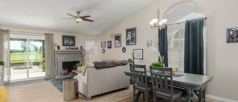 A spacious living room with a fireplace, a dining area with a table and chairs, and a ceiling fan.