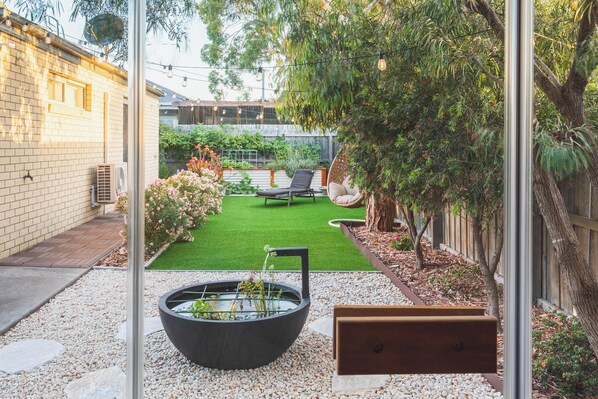 Un patio trasero con un césped pequeño, una jardinera negra con plantas, un banco de madera y una silla colgante.