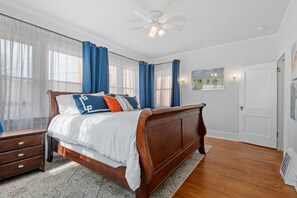 Un dormitorio con cama de madera, ropa de cama blanca, cortinas azules, ventilador de techo y un cuadro en la pared.