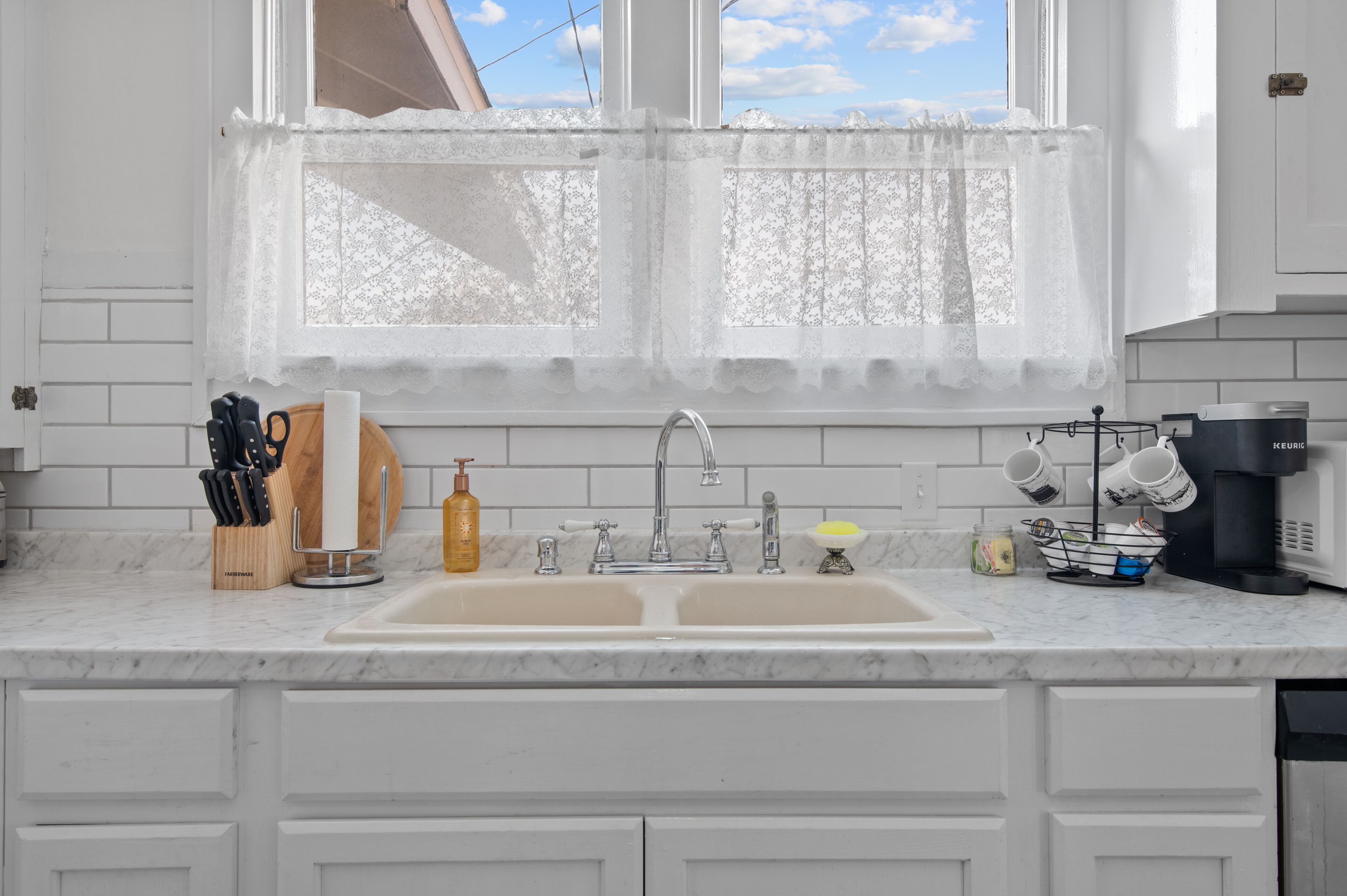 A kitchen with a double sink, white cabinets, a window with lace curtains, and various kitchen utensils and appliances.