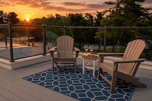 Une terrasse avec deux fauteuils Adirondack, une petite table sur laquelle se trouve un plateau de nourriture et deux verres de vin, et une vue sur le coucher de soleil au-dessus d’une zone boisée.