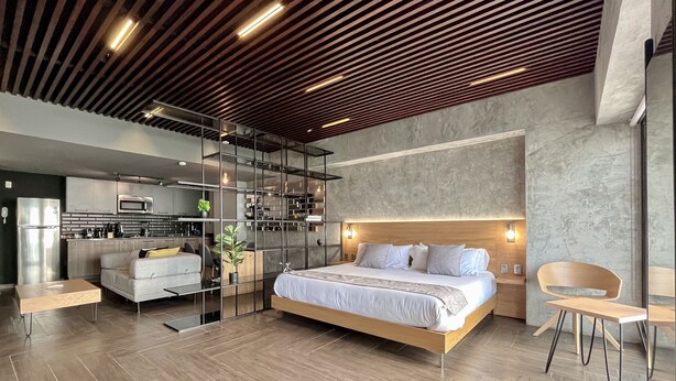 Un dormitorio moderno con una cama grande, techo de madera y una cocina visible a través de un panel de vidrio.