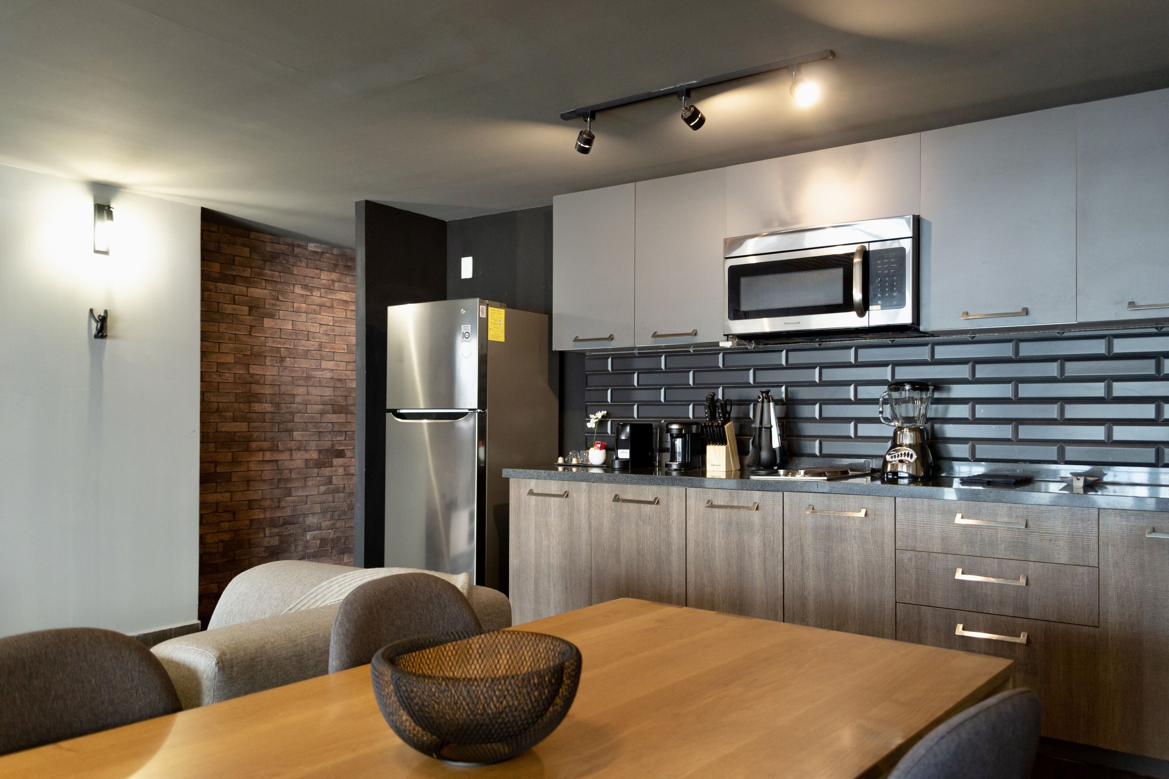 A modern kitchen with a stainless steel refrigerator, a microwave, and a coffee maker. There is a brick accent wall and a wooden dining table with chairs.