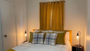 Un lit bien fait, avec une couette blanche, un plaid jaune et plusieurs oreillers. On peut voir deux tables de chevet avec des lampes. Une porte est visible sur le côté gauche de l’image.