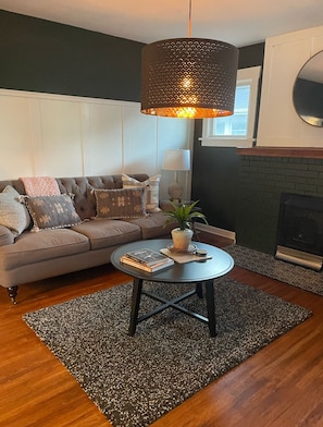 A living room with a dark green wall, a brown sofa with patterned cushions, a round coffee table with books and a plant, a hanging lamp, and a fireplace.