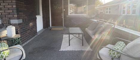 A covered patio area with a brick wall, a glass door, and a seating arrangement including a table and chairs.