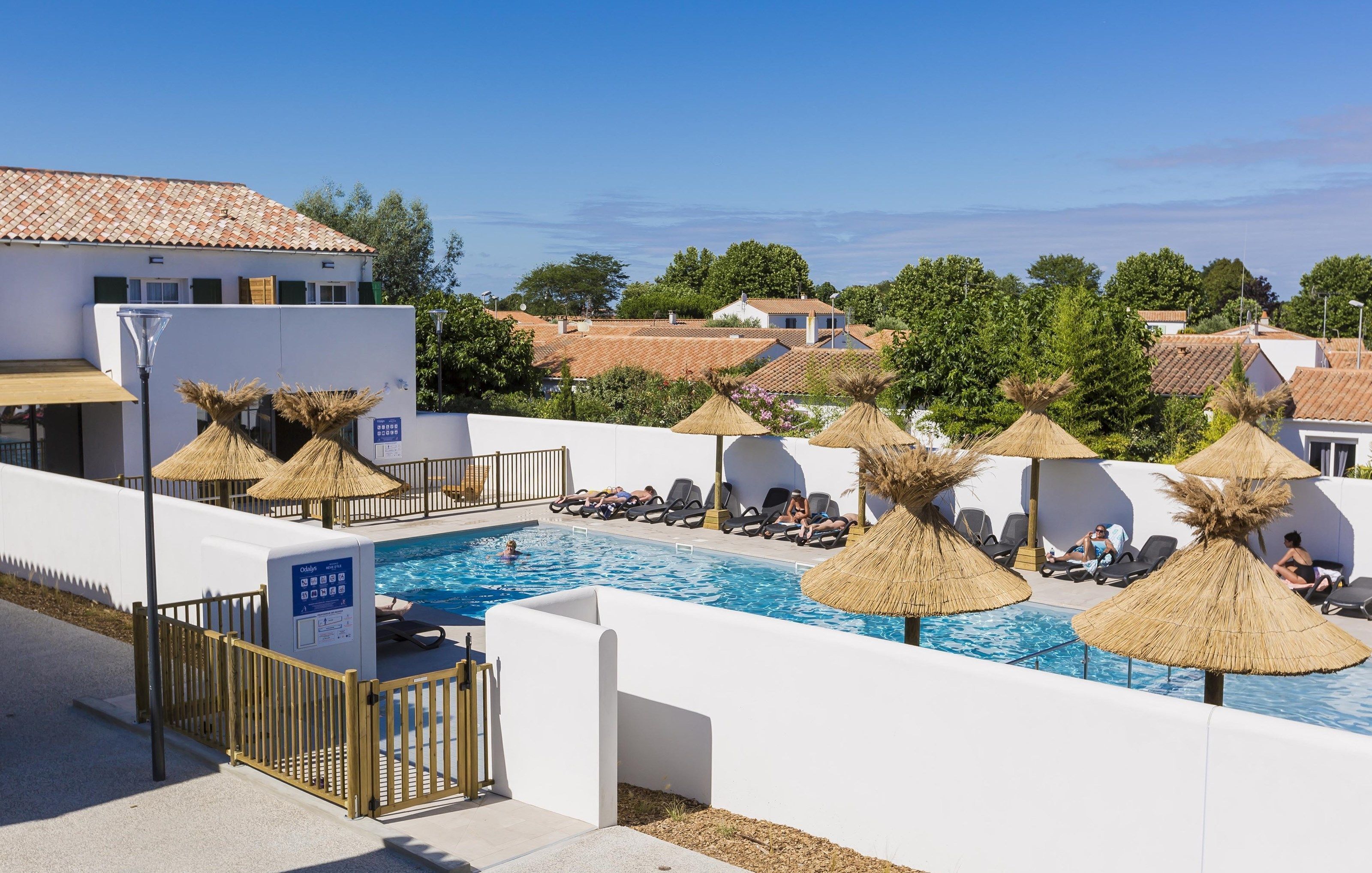 A swimming pool with sun loungers and thatched umbrellas, surrounded by white walls and a fence.