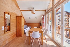 A wooden interior with a dining table and chairs, large windows, and a view of a building outside.