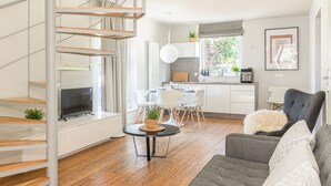 Een moderne leefruimte met een spiltrap, een eethoek met witte stoelen, een grijze bank en een keuken met een koffieapparaat.