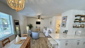Eine moderne Küche mit weißen Schränken, ein Essbereich mit Holztisch und -stühle sowie ein Wohnzimmer mit Sofa und Deckenventilator.