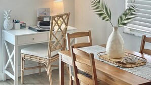 Ein Essbereich mit einem weißen Tisch, Holzstühlen, einem Korbstuhl, einer weißen Vase mit Blättern und einer Kandelaber.