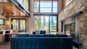 Una sala de estar moderna con un sofá modular grande, una chimenea y ventanas amplias con vistas a árboles y montañas.