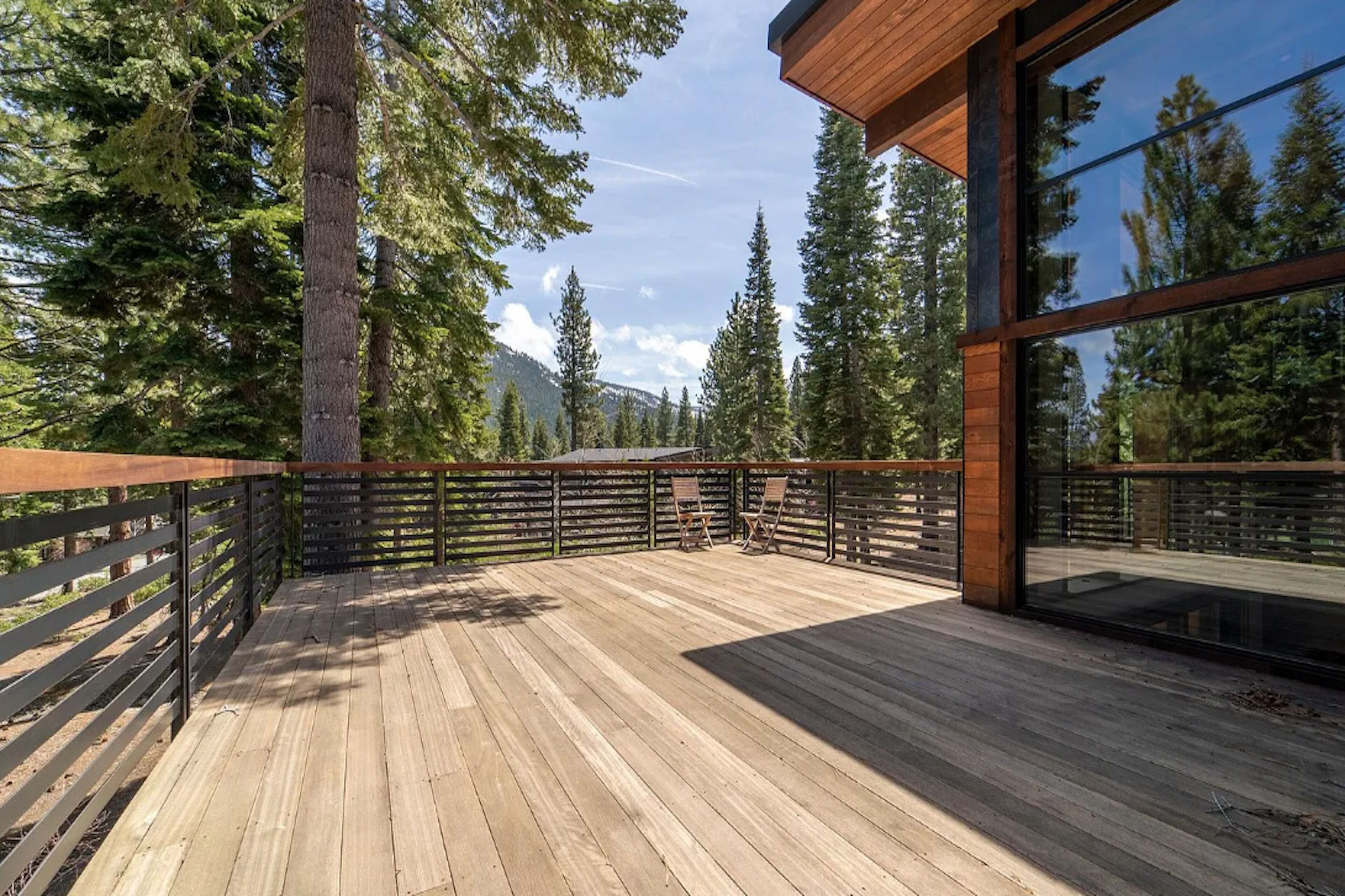 Una terraza de madera con cerramiento de vidrio, rodeada de altos árboles y montañas.