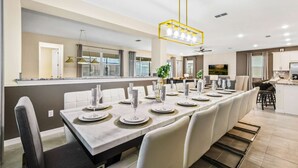 Een moderne eetruimte met een lange tafel voor zes personen, witte stoelen en een centrale kroonluchter.