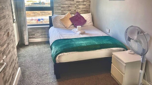 A bedroom with a bed, a window with a view, a fan, and a wall-mounted picture. - Remo Apartment Near Canary Wharf & 02 (London)