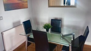 A modern dining area with a glass table, black chairs, a potted plant, a framed mirror, and a sunset painting on the wall. - Remo Apartment Near Canary Wharf & 02 (London)