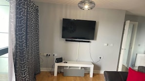 A modern living room with a flat-screen TV mounted on the wall, a white entertainment unit, a laptop on a small table, and a sofa with a red cushion. - Remo Apartment Near Canary Wharf & 02 (London)
