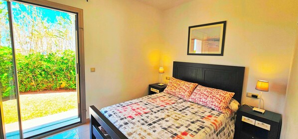 Ein Schlafzimmer mit einem Bett, zwei Nachttischen mit Lampen, einem gerahmten Bild an der Wand und einer Glastür, die in einen Garten führt.