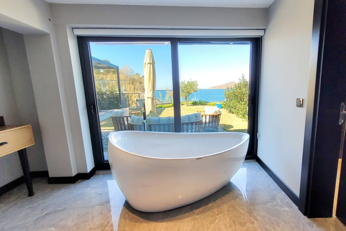 Un baño moderno con una bañera independiente, un lavamanos y un amplio ventanal corredero que da a un patio exterior con vistas al mar.