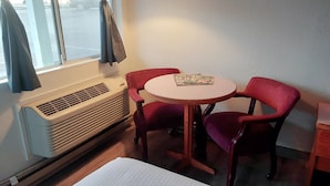 Ein Hotelzimmer mit rundem Tisch, zwei roten Stühlen, einem Fenster mit Vorhängen, einer Klimaanlage und einem kleinen Nachttisch mit Lampe.