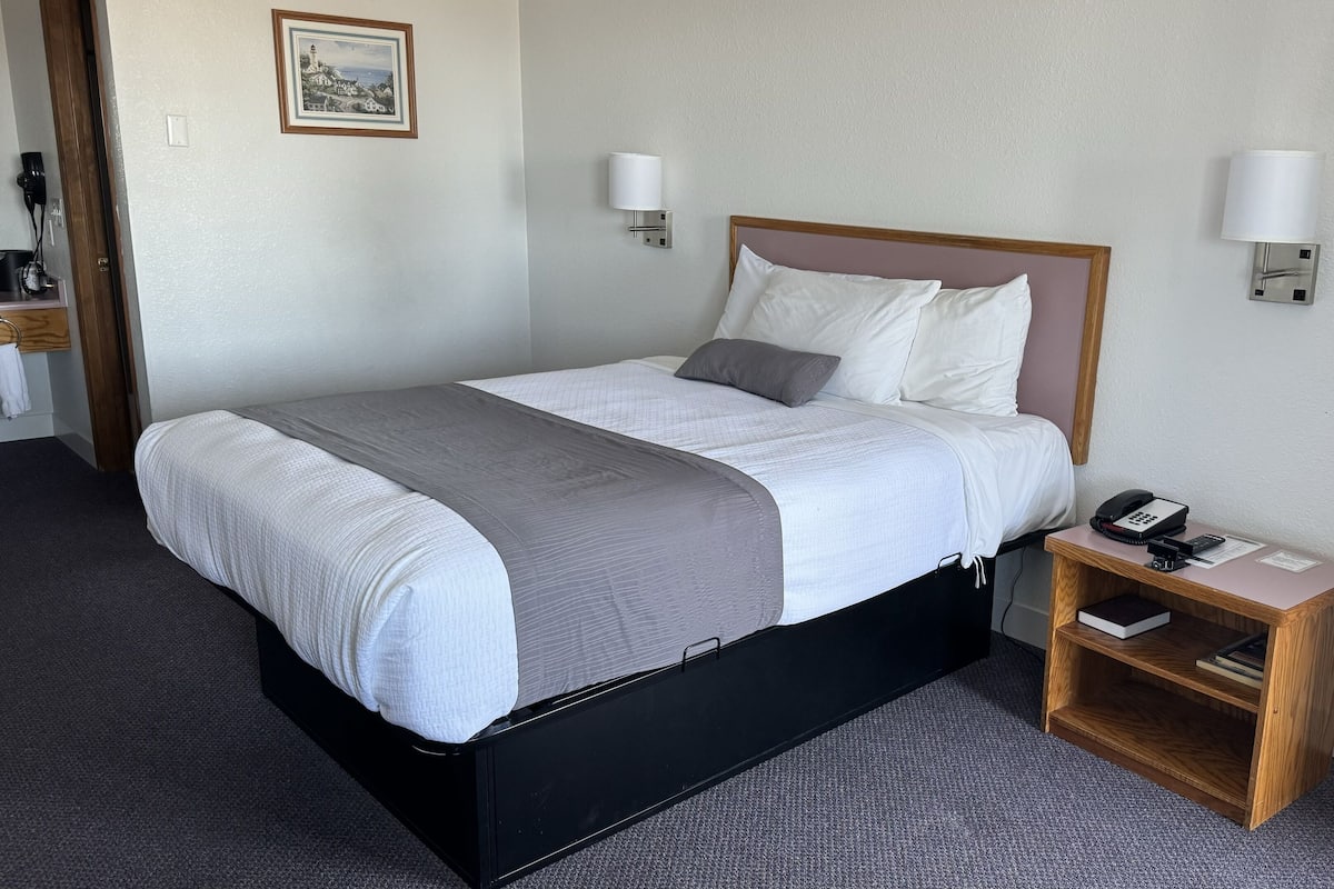 Una cama bien hecha con una colcha gris y blanca, un cabecero de madera, dos lámparas de noche, un teléfono junto a la cama y una mesita de noche de madera con un libro y un control remoto.
