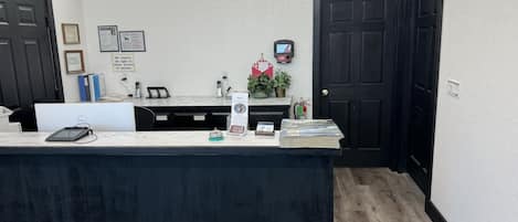 A reception desk with a computer, phone, and informational pamphlets. There are framed documents on the wall, a small plant, and a fire extinguisher.