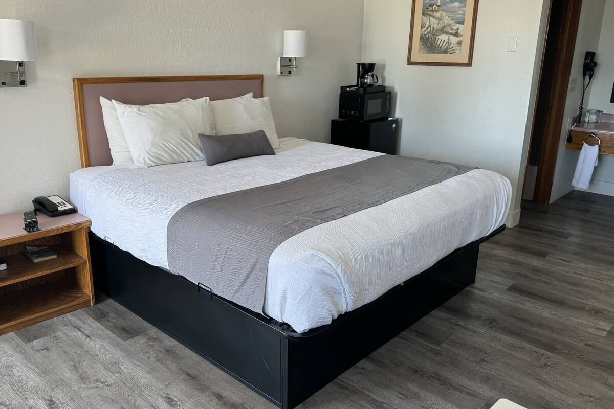 Una cama bien hecha con un edredón gris y blanco, una mesita de noche con un teléfono y un libro, y una lámpara fijada a la pared.