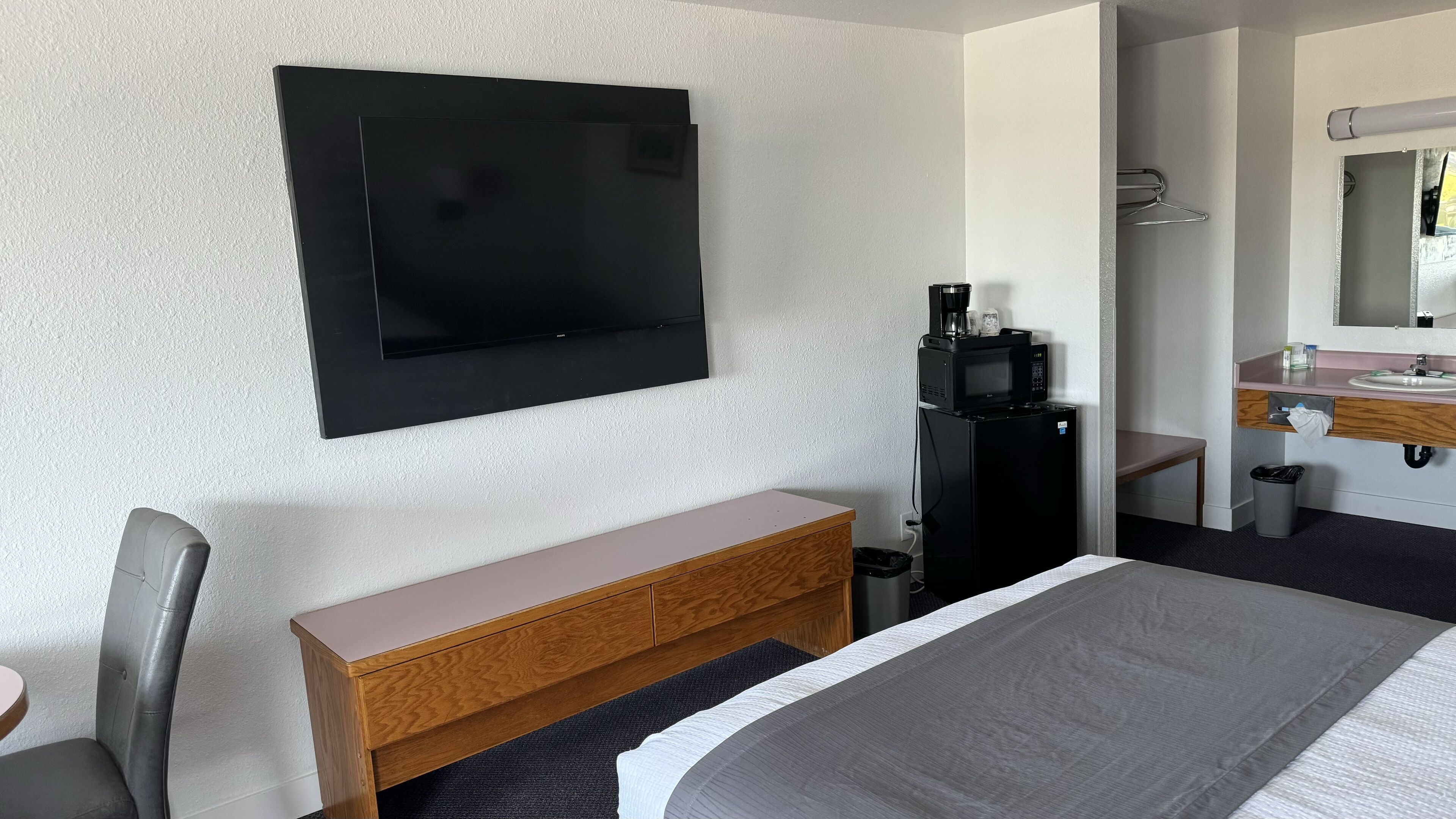 Quarto de hotel com cama, mesa de cabeceira de madeira, escrivaninha, cadeira, televisão na parede, micro-ondas, lixeira e banheiro visível por uma porta aberta.