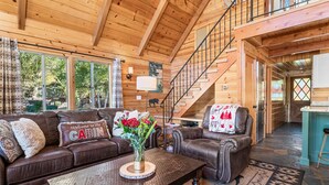 A cabin interior with a wooden ceiling, a staircase, a living room with a sofa and armchairs, and a dining area.
