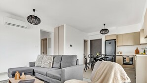 Un appartement moderne avec un canapé gris, une table basse en bois, un coin repas avec une table et des chaises noires, et une cuisine équipée d’un réfrigérateur et d’un four.
