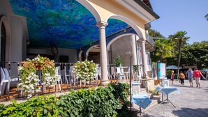 An outdoor seating area with white chairs and tables, surrounded by greenery and a blue mural on the ceiling.