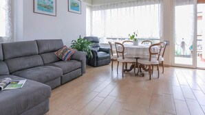 A living room with a grey sofa, a dining table with chairs, and a large window with sheer curtains.